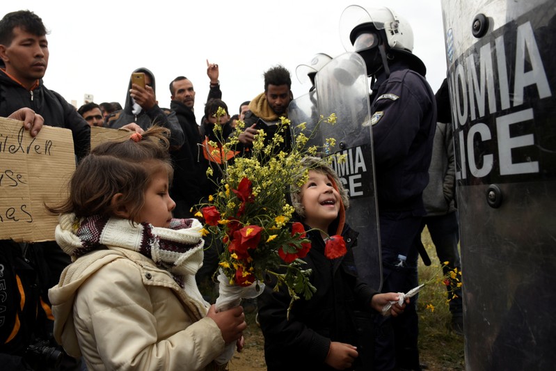 Migrant children hold flowers as they stand in front of riot police officers next to a camp in the town of Diavata in northern Greece