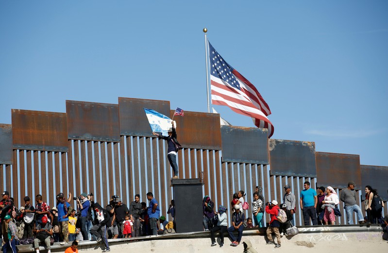 FILE PHOTO: A migrant, part of a caravan of thousands traveling from Central America en route to the United States, holds flags of Honduras and the United States in front of the border wall between the U.S. and Mexico in Tijuana