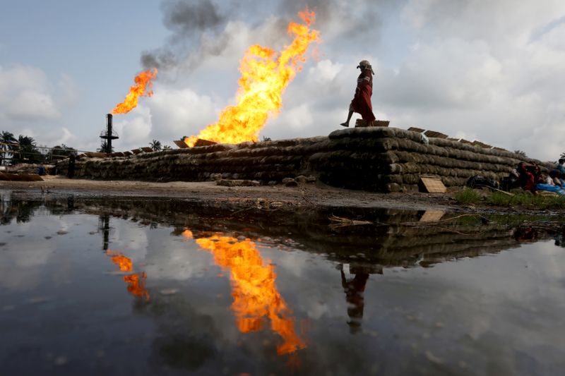 FILE PHOTO: A reflection of two gas flaring furnaces and a woman walking on sand barriers is seen in the pool of oil-smeared water at a flow station in Ughelli, Delta State