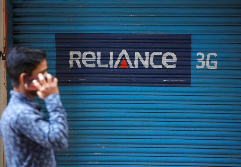 FILE PHOTO: A man speaks on his mobile phone as he walks past a closed shop painted with an advertisement of Reliance Communications in Mumbai