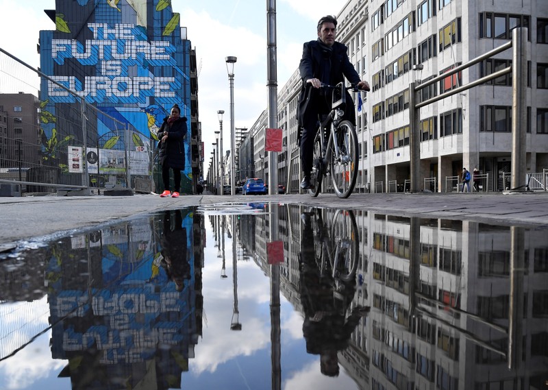 People are reflected in a puddle as they pass by a mural near the EU headquarters in Brussels