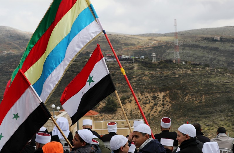 Members of the Druze community holds Syrian and Druze flags as they sit facing Syria, during a rally marking the anniversary of Israel's annexation of the Golan Heights in the Druze village of Majdal Shams, in the Israeli-occupied Golan Heights